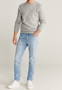 Suéter de algodón gris con mangas largas, combinado con jeans de un azul claro y zapatillas blancas. Los jeans tienen un aspecto desgastado y un diseño clásico de cinco bolsillos.