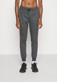 Hombre vistiendo pantalones deportivos gris oscuro con franjas rojas a los lados, zapatillas negras y calcetines blancos, de pie frente a un fondo claro y liso.