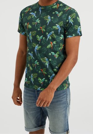 Mężczyzna w ciemnozielonej koszuli z krótkim rękawem, ozdobionej kolorowym wzorem tropikalnych papug i liści, w połączeniu z podwiniętymi dżinsowymi szortami.