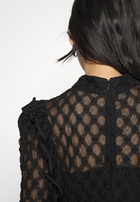 Frau von hinten gesehen, die ein schwarzes Spitzentop mit Blumenmustern, Rüschen an den Schultern und einem kleinen Reißverschluss auf der Rückseite trägt.