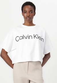 Hvit avkortet t-skjorte laget av glatt bomull med korte ermer. Har en markant svart "Calvin Klein"-logo foran. Enkel design.