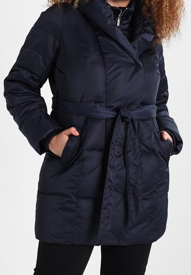 Personne portant un manteau matelassé bleu marine avec une ceinture nouée à la taille et les mains dans les poches latérales, visible des épaules aux cuisses.