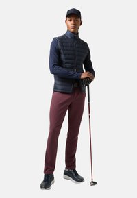 Pánský golfový outfit: námořnická prošívaná vesta, dlouhý rukáv, kaštanové kalhoty, černé tenisky. Drží golfovou hůl. Jednoduchá kšiltovka.