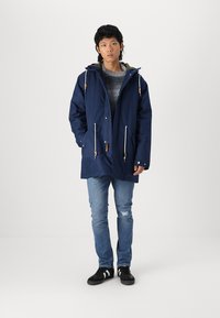 Marineblauwe waterdichte parka met een capuchon, voorzien van trekkoorden, voorkeurszakken en een grijze voering, gecombineerd met lichtblauwe jeans en zwarte sneakers.