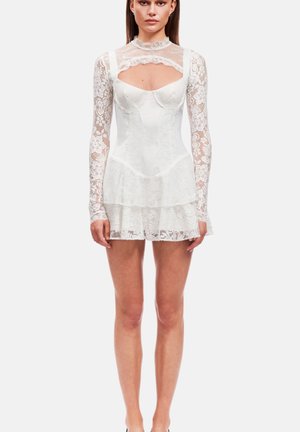 Woman wearing a white lace mini dress with long sheer sleeves, high neckline, and tiered skirt, standing against a plain background.