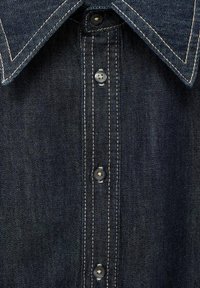Chemise en denim bleu foncé avec un col pointu, patte de boutonnage avec des boutons en métal et des coutures beiges contrastantes le long des coutures.