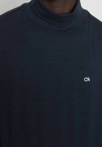 Mörkblåt polotröja i mjukt tyg, med en figurnära design och en liten vit "ck"-logotyp på bröstet. Slät struktur, minimalistisk stil.