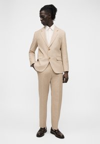 Jeune homme portant un costume beige à deux boutons avec un pantalon assorti, une chemise blanche et des mocassins brun foncé, debout avec une main dans la poche.