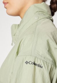 Vaaleanvihreä kevyt takki, jossa on korkea kaulus, painonapit ja kirjailtu "Columbia" -logo vasemmassa olkapäässä. Sileä pinta.