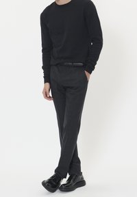 Mies, který má na sobě černou dlouhounkou košili, tmavě šedé vzorované kalhoty, černý kožený opasek a lesklé černé formální boty, stojí s jednou rukou v kapse.