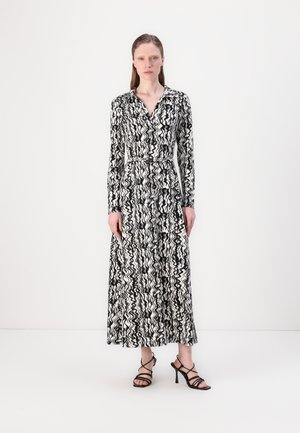 Robe longue à manches longues noir et blanc avec un motif ondulé, design avec col, taille ceinturée et jupe longue. Portée avec des sandales à talons noires à lanières.