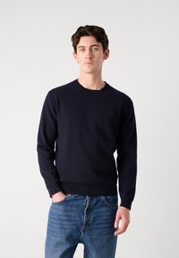 Navy knitted sweater with a round neckline and ribbed cuffs. Features a subtle logo on the chest. Paired with blue denim jeans.