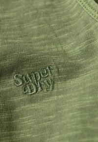 Tkanina w oliwkowym kolorze z miękkim, teksturowanym wykończeniem, z haftowanym logo "Superdry" w nieco ciemniejszej zielonej nici.