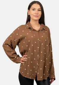Brown button-up shirt in textured fabric with white heart patterns, featuring a collar and long sleeves, worn over black leggings.