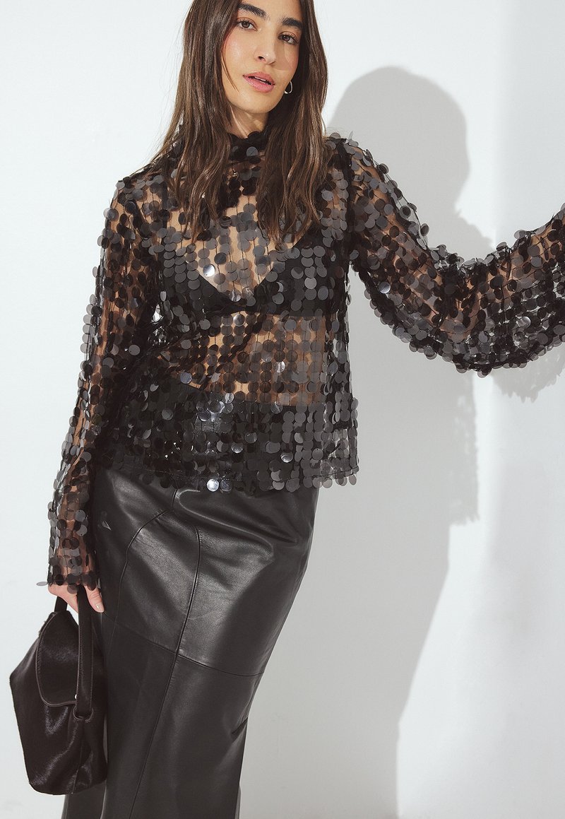 Mustat läpikuultavatätö, jossa on pyöreitä paljettikoristeita ja pitkät hihat, yhdistettynä mustaan nahkahameeseen ja pieneen mustaan käsilaukkuun.