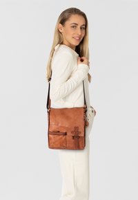 Borsa a tracolla in pelle marrone con superficie liscia, tracolla regolabile, tasca frontale e dettaglio in metallo decorativo.