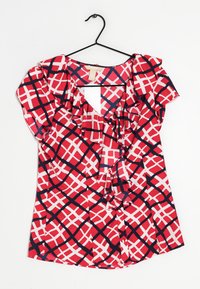 Blusa rossa e bianca con scollatura arricciata, caratterizzata da un motivo a incrocio rifinito in blu navy. Il tessuto leggero pende da un appendiabiti nero.
