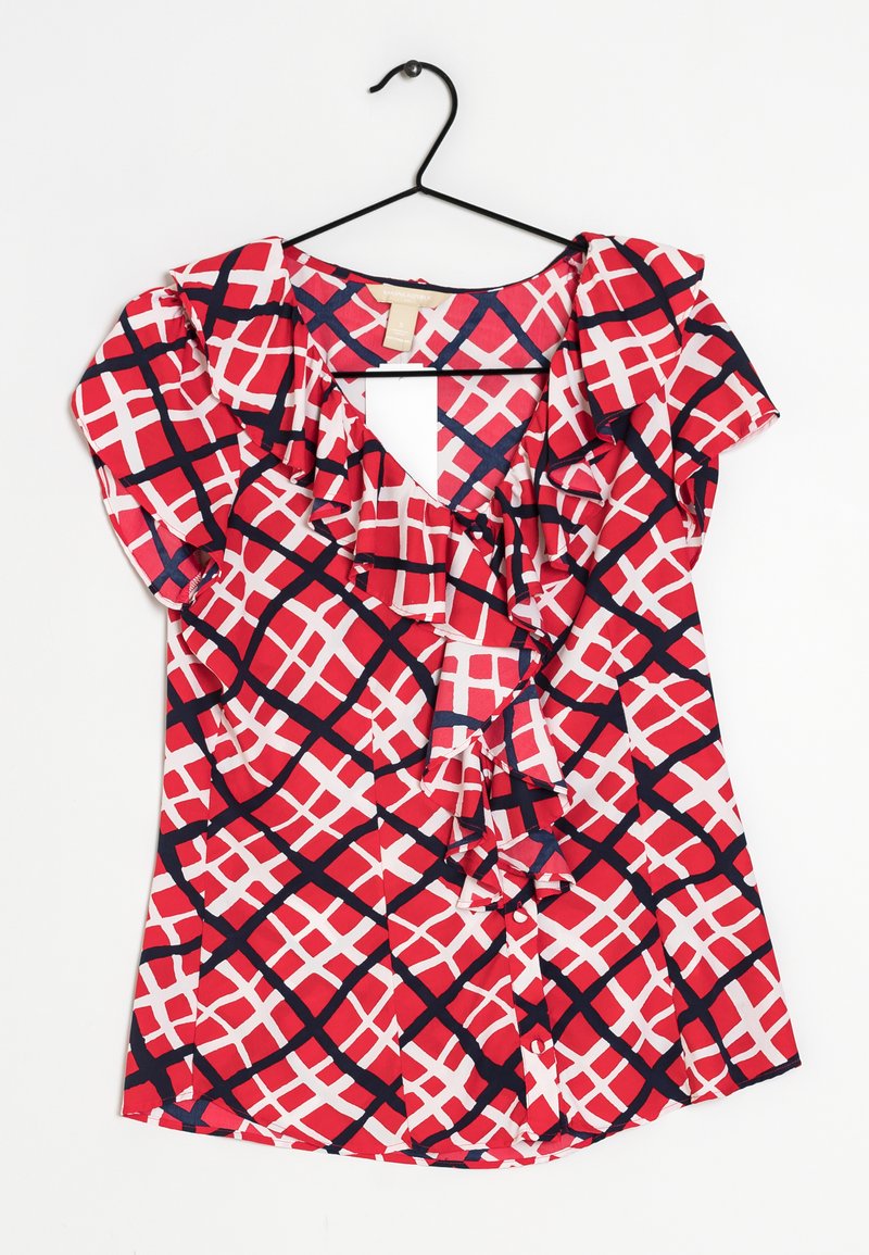 Blusa rossa e bianca con scollatura arricciata, caratterizzata da un motivo a incrocio rifinito in blu navy. Il tessuto leggero pende da un appendiabiti nero.