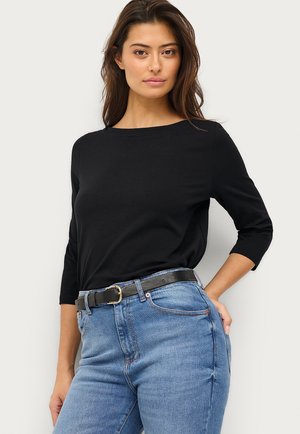 Junge Frau in einem schwarzen Oberteil mit drei Viertel Ärmeln, das in hochtaillierte blaue Jeans mit einem schwarzen Gürtel gesteckt ist, steht vor einem weißen Hintergrund.