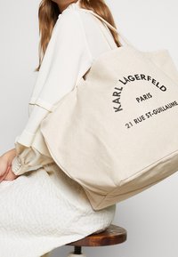 Woman in cream blouse and skirt sits on wooden stool, carrying beige Karl Lagerfeld tote bag with black text and handles over shoulder.