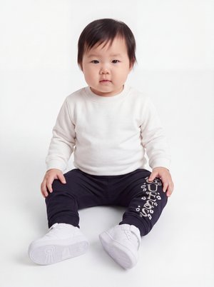 Kleinkind mit dunklem Haar sitzt, trägt ein weißes Langarmshirt, schwarze Hose mit weißem Text und weiße Turnschuhe auf weißem Hintergrund.