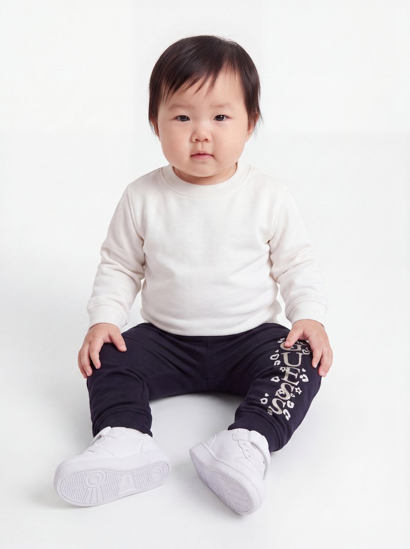 Tout-petit aux cheveux foncés assis, portant un tee-shirt blanc à manches longues, un pantalon noir avec texte blanc et des baskets blanches sur fond blanc.