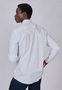 Chemise blanche à pois noirs, à manches longues, avec un pli au dos. Texture lisse, ourlet droit et poignets à boutons.