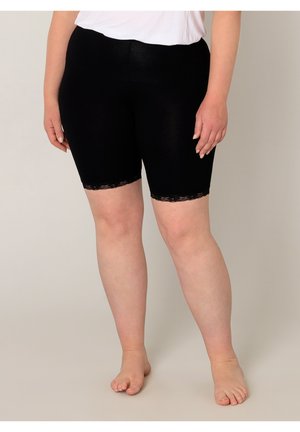 Persoon draagt zwarte knielange leggings met kanten randje, staat blootsvoets tegen een effen beige achtergrond.