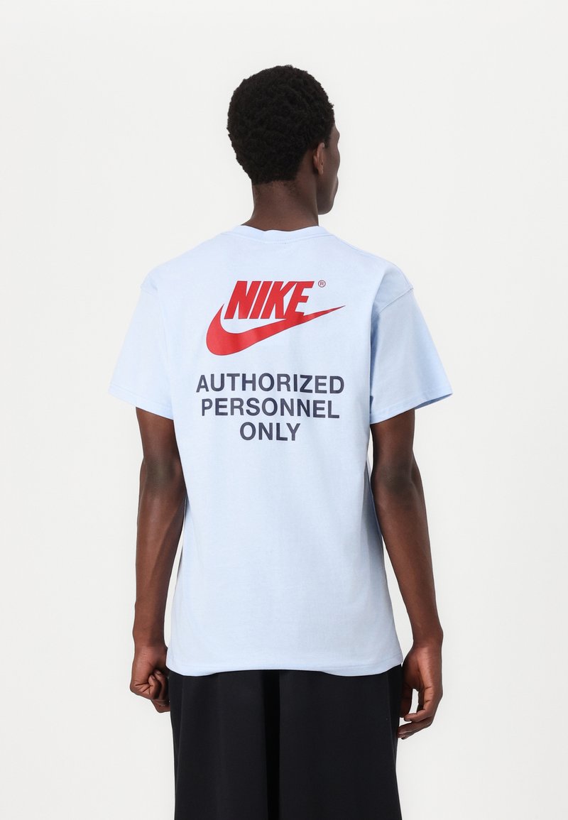 Gaiši zils kokvilnas T krekls ar īsām piedurknēm, kurā aizmugurē ir sarkans Nike logotips un melns teksts "TIKAI AUTORIZĒTAM PERSONĀLAM".