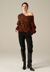 Maglione marrone lavorato a maglia con design fuori spalla e orlo a coste, abbinato a pantaloni neri e stivali lucidi al ginocchio.