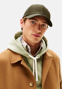 Gorra de oliva, abrigo de lana marrón y sudadera verde claro en capas sobre una camisa de cuadros. Presenta un cordón blanco y detalles de botones.