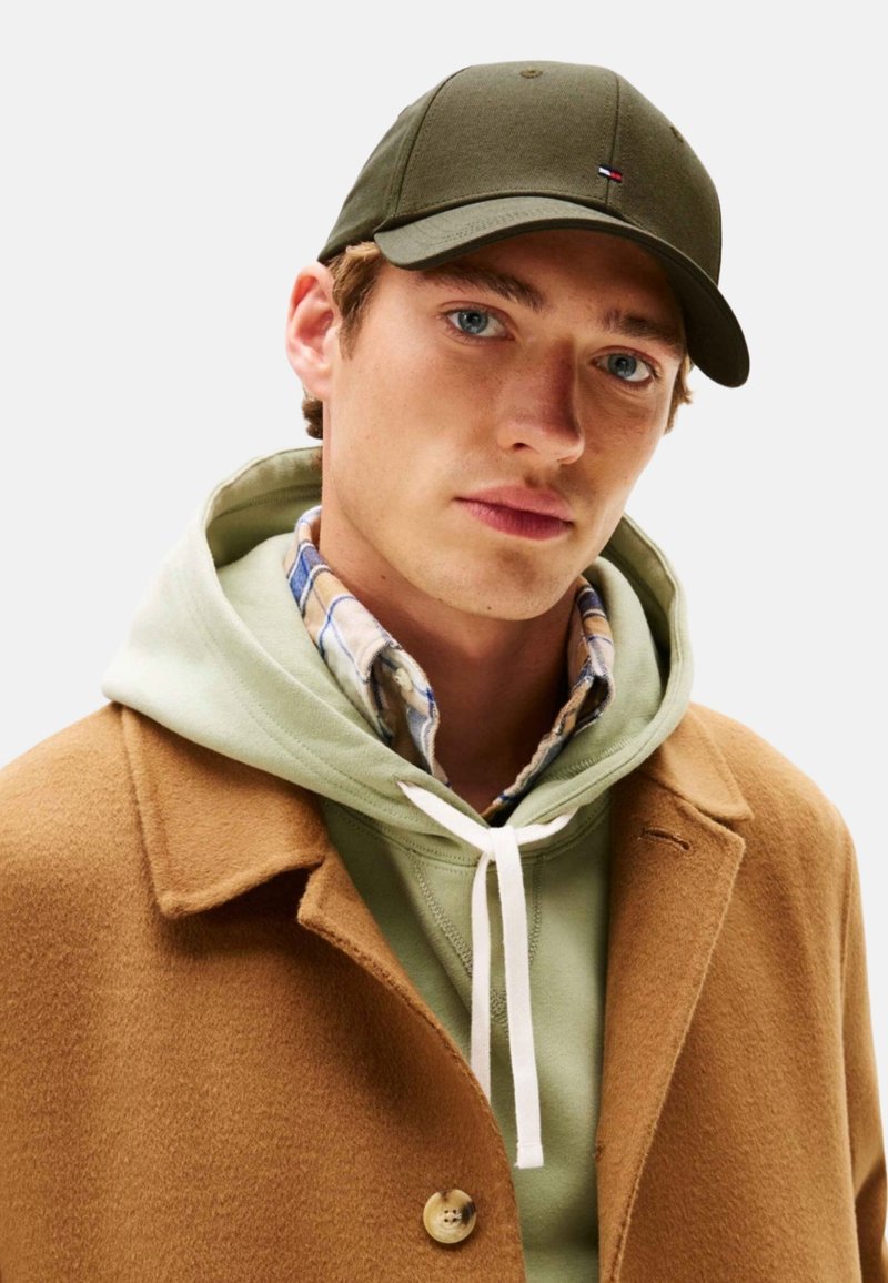 Gorra de oliva, abrigo de lana marrón y sudadera verde claro en capas sobre una camisa de cuadros. Presenta un cordón blanco y detalles de botones.