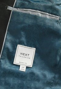 Funda para ropa de color azul con un diseño estampado y una textura suave, que incluye un bolsillo con cremallera plateada y una etiqueta blanca con la inscripción "NEXT TAILORING SKINNY FIT".