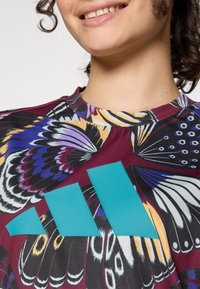 Tissu multicolore avec un motif de papillons ; logo Adidas turquoise sur fond bordeaux ; design à manches courtes avec un col rond.