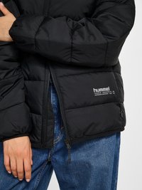 Osoba w czarnej puchowej kurtce z widocznym logo hummel oraz w niebieskich dżinsach, z założonymi rękami i częściowo rozpiętą kurtką.