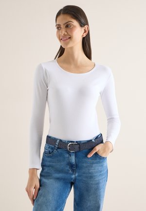 Vrouw draagt een nauwsluitende witte top met lange mouwen en blauwe spijkerbroek met een donkere riem met patroon, staand met één hand in haar zak.