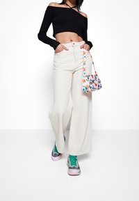 Weiße weite Hose, schwarzes schulterfreies geripptes Croptop, farbenfrohe gemusterte Handtasche und grüne sowie rosa Sneakers.