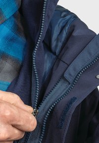 Marinejacke aus strukturiertem Stoff, mit blauem Reißverschluss und Innenfutter. Hand, die den Reißverschlussgriff zum Schließen hält.