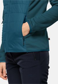 Veste isolante bleu sarcelle avec une texture lisse, des poches zippées et un design à panneaux contrastés. Associée à un pantalon bleu marine foncé avec une fermeture éclair de poche.