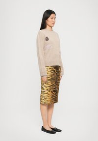 Maglione beige lavorato a maglia con dettagli ricamati colorati, abbinato a una gonna a strisce tigre in oro e nero lunghezza ginocchio, e ballerine nere.