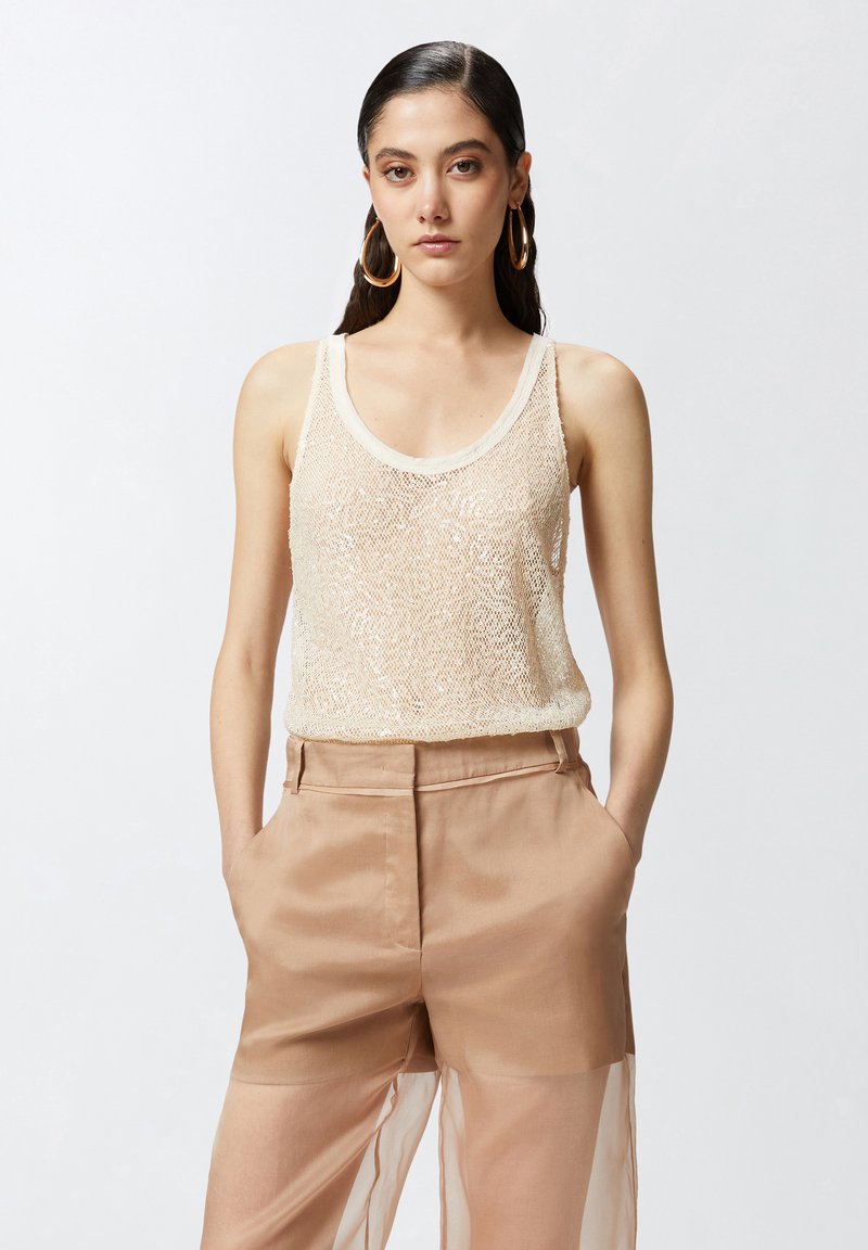 Bež majica bez rukava ukrašena perlicama s izrezanim vratnim otvorom, u kombinaciji s svijetlosmeškim, prozračnim hlačama širokih nogavica koje imaju bočne džepove i glatku teksturu.