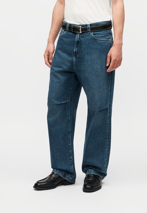 Άντρας που φοράει άνετο μπλε τζιν, μαύρη δερμάτινη ζώνη, άσπρο πουκάμισο και μαύρα δερμάτινα παπούτσια, στέκεται μπροστά σε απλό φόντο.