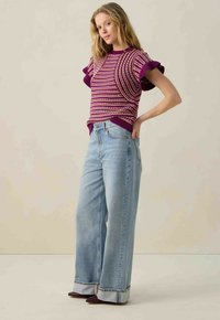 Top en tricot avec une base violette, motif rayé en or et crème ; associé à un jean à jambes larges bleu clair avec des poignets retroussés et des chaussures foncées.