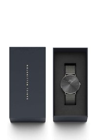 Black mesh strap watch in a navy blue box with gold text. Round gray dial features silver marks and logo, resting on a soft black interior.
