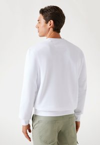 White, long-sleeve sweatshirt with ribbed cuffs and hem. Soft fabric, minimalist design, featuring a small back detail. Worn with light green pants.