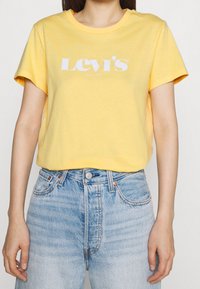 Žluté bavlněné tričko s krátkým rukávem a kulatým výstřihem, s bílým logem "Levi's" potištěným na hrudi. Nosené s modrými džíny.