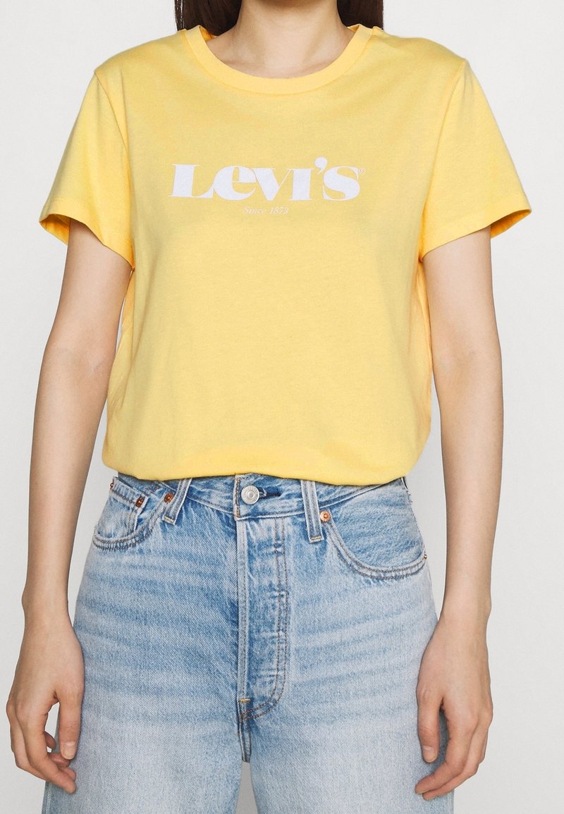 Žluté bavlněné tričko s krátkým rukávem a kulatým výstřihem, s bílým logem "Levi's" potištěným na hrudi. Nosené s modrými džíny.