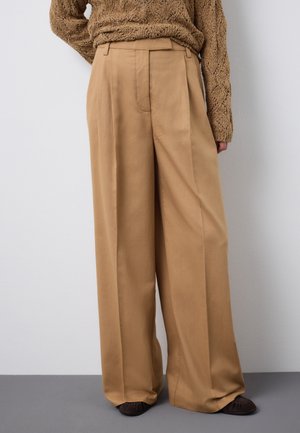 Beige Hosen mit weitem Bein, kombiniert mit einem strukturierten braunen Pullover und dunkelbraunen flachen Schuhen, vor einem neutralen Hintergrund stehend.