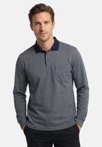 Mann mit den Händen in den Taschen, der ein graues lang ärmeliges Poloshirt mit einem dunklen Kragen und einer Brusttasche vor einem einfarbigen Hintergrund trägt.