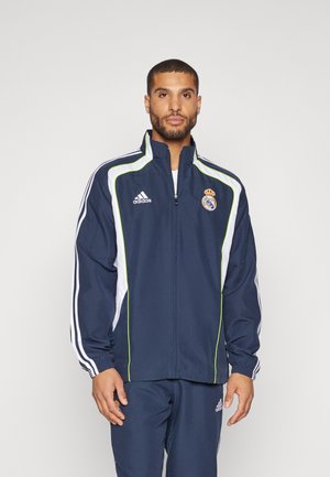 Navy athletic jacket with white and green accents, full zip, high collar, and Real Madrid logo. Made of lightweight material.